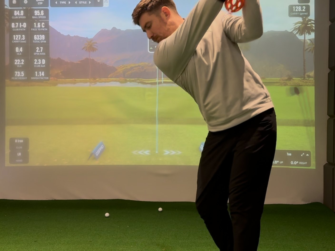 Golfer mid-swing in the indoor studio with live launch monitor data on screen